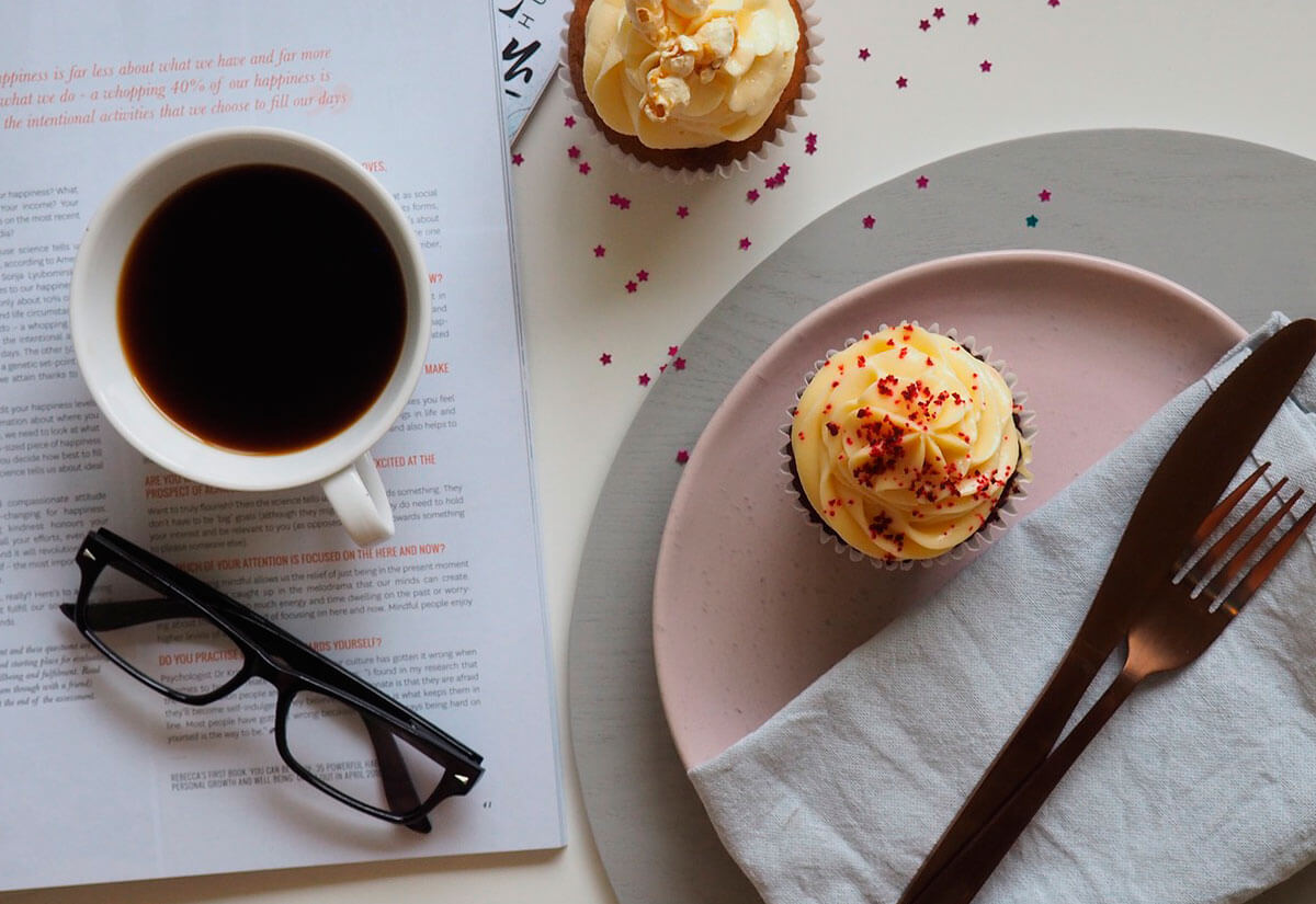 Imagen de una taza café con cubiertos, postres, unas gafas y platos